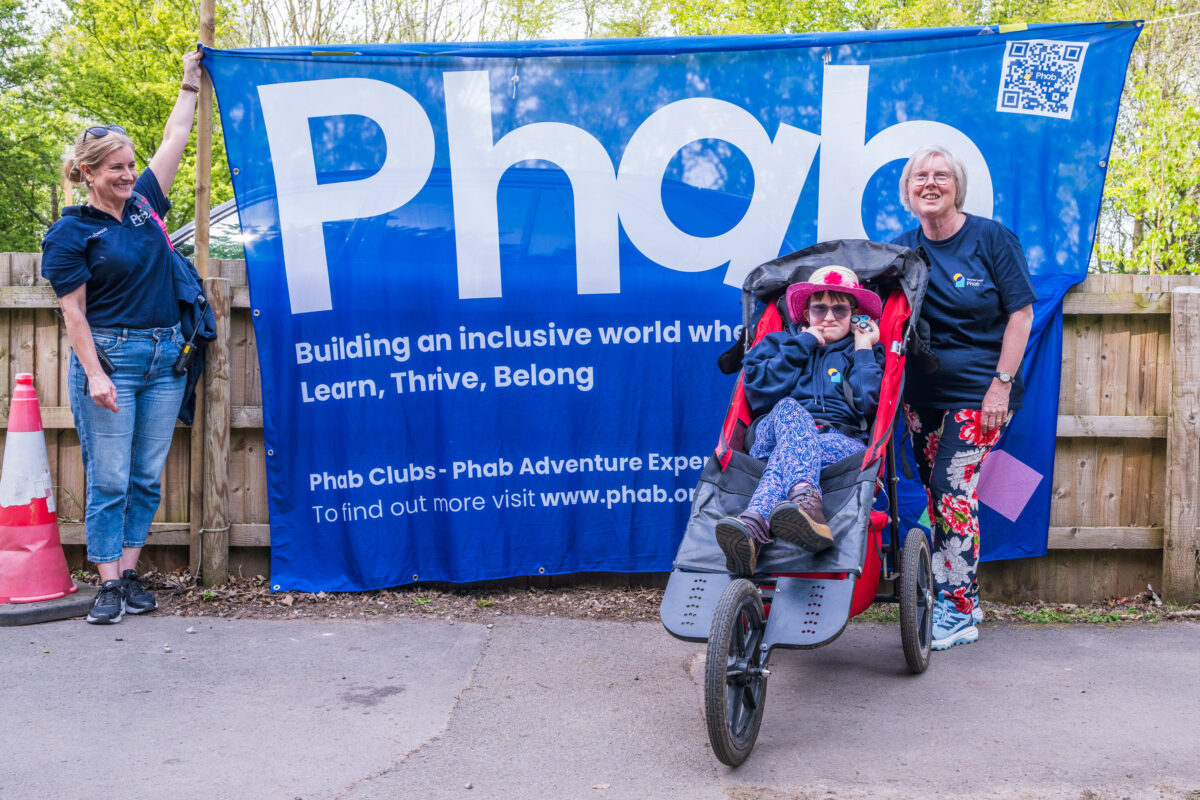 Three people stand in front of a large blue Phab banner at an outdoor event, with one person seated in an adapted wheelchair in the centre and two others standing on either side, on a paved path bordered by wooden fencing and trees.