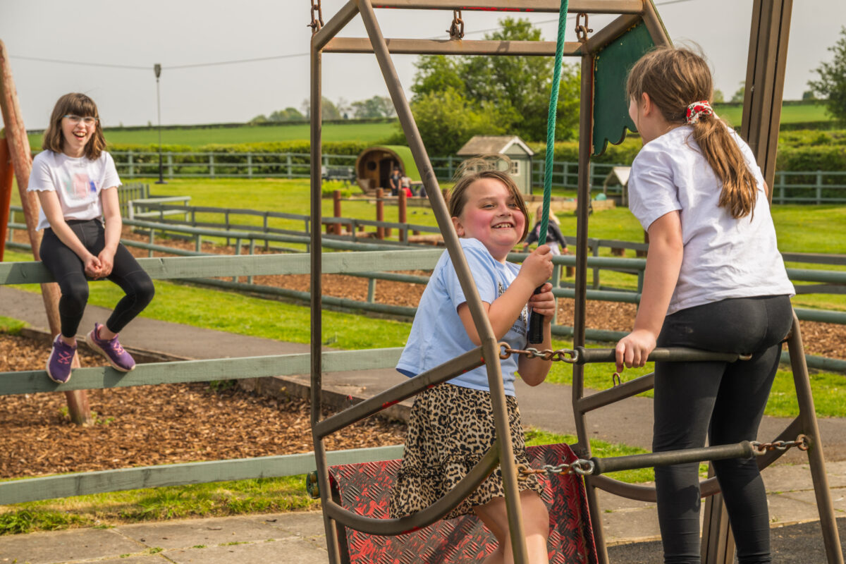 Three children play together on an outdoor play structure, with one child standing inside a metal climbing frame, another holding onto a rope, and a third sitting nearby on a wooden fence, with grassy fields and fenced pathways in the background.