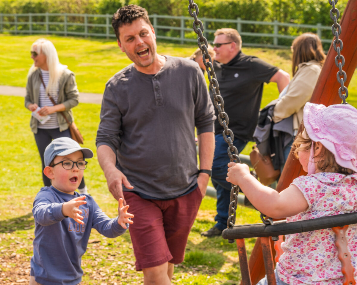 A child wearing glasses reaches out toward another child sitting on a playground swing, while an adult stands nearby supervising, with other adults and a grassy outdoor area in the background.