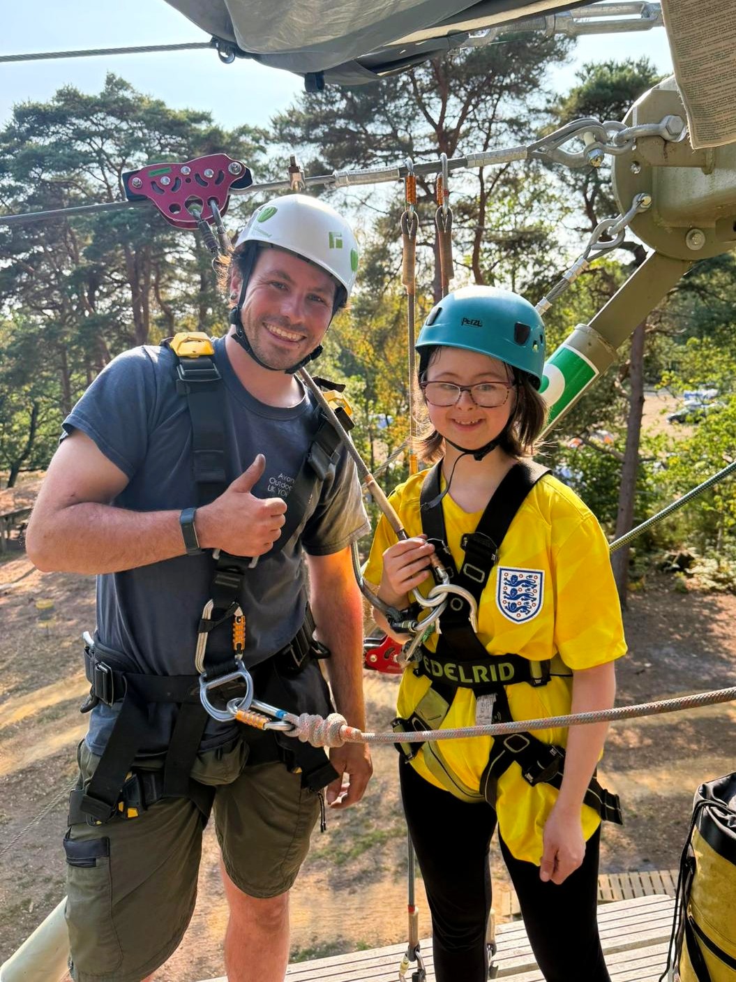Two people wearing safety helmets and harnesses stand on a wooden platform at a high ropes course, clipped into cables, with trees and ropes visible in the background.