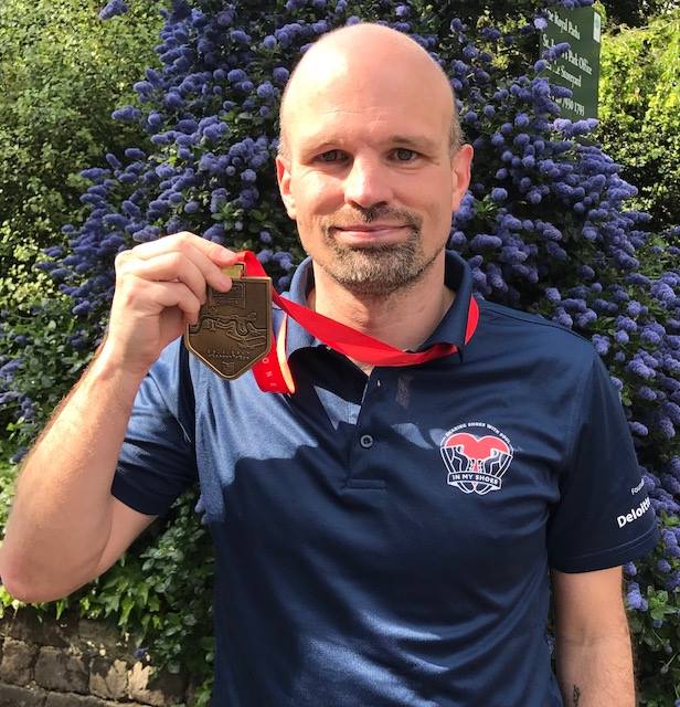 A person stands outdoors in front of a large bush covered in purple flowers, holding up a bronze-colored medal on a red ribbon. The person is wearing a navy polo shirt with a “In My Shoes” logo on the chest. Sunlight highlights the scene, and greenery fills the background.