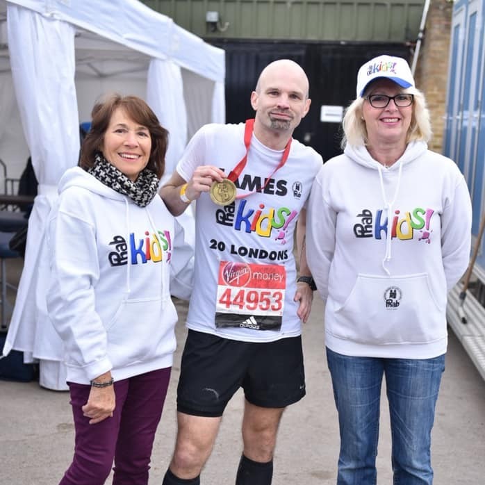Three people stand together at an outdoor event near white tents. The person in the middle wears a white “Phab Kids” T-shirt, a marathon race bib numbered 44953, and black running shorts, holding up a gold medal on a red ribbon. The two people on either side wear matching grey “Phab Kids” hoodies, with one also wearing a white cap. The group is positioned on a paved area with event structures and equipment visible in the background.