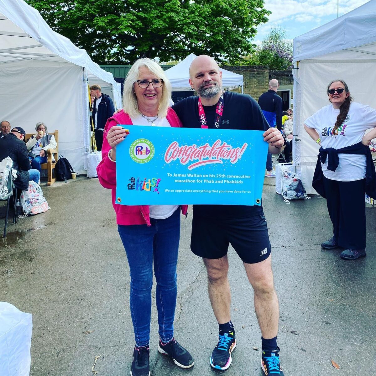 Two people stand outdoors between white tents at an event, holding a bright blue banner that reads: “Congratulations! To James Walton on his 25th consecutive marathon for Phab and PhabKids. We so appreciate everything that you have done for us.” One person is wearing running gear and a medal, and several other people, chairs, and bags are visible around the event space. A large green tree and blue sky appear in the background.