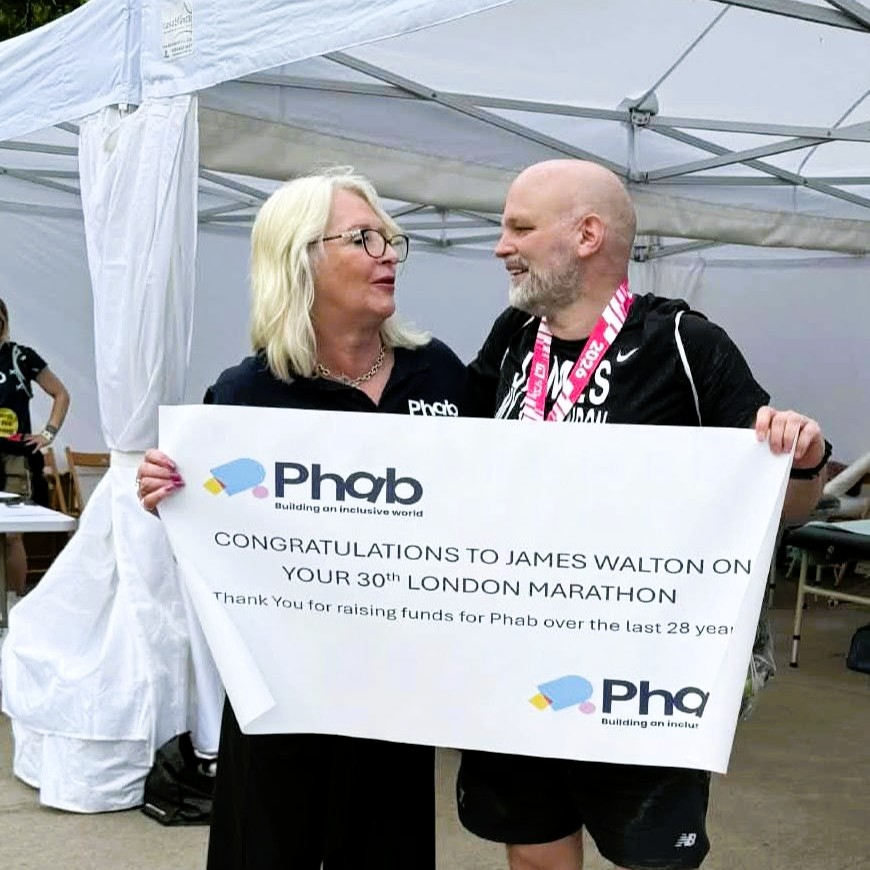 Two people stand under a white event tent holding a large printed sign together. The sign reads: “Phab – Building an inclusive world. Congratulations to James Walton on your 30th London Marathon. Thank you for raising funds for Phab over the last 28 years.” One person is wearing a medal, and tables and event equipment are visible in the background.