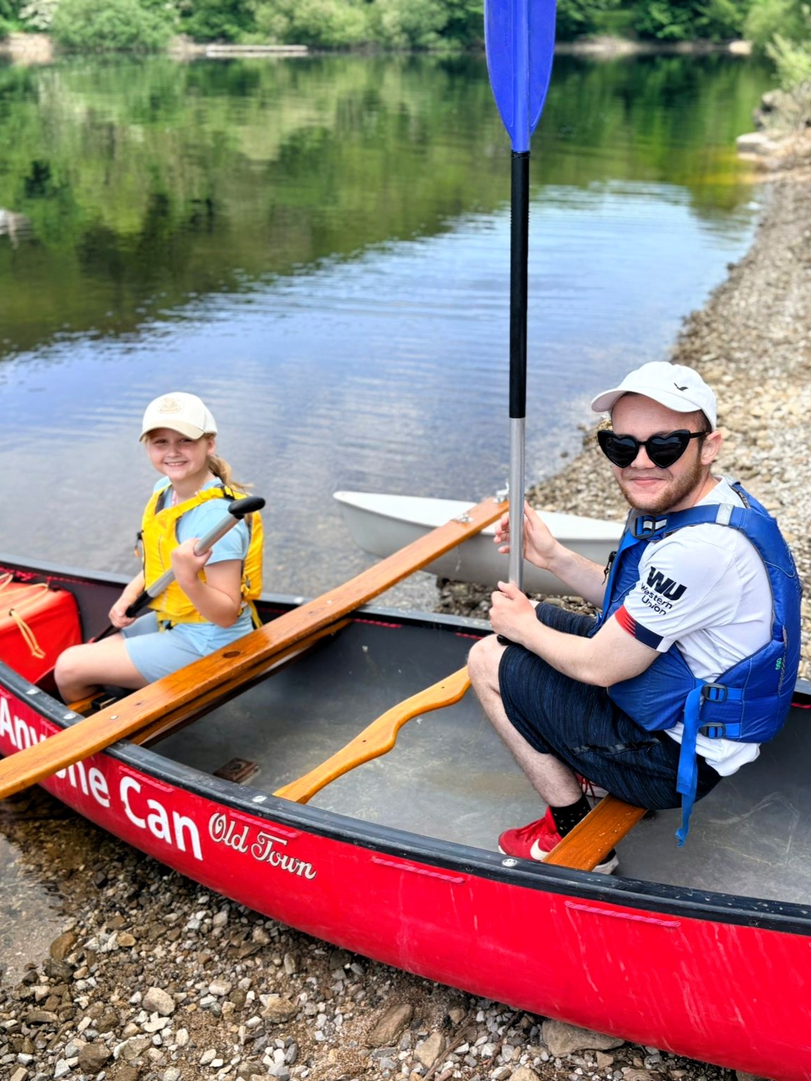 Two people wearing life jackets sit in a red canoe at the edge of a lake, each holding a paddle. The canoe rests on a pebbled shoreline with calm water and green trees in the background.