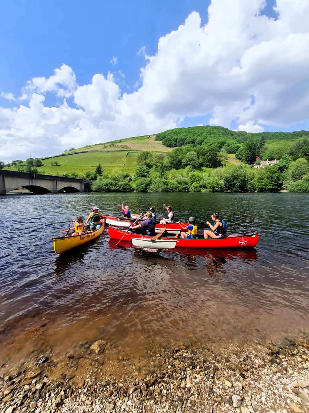 A group of people paddle red and yellow canoes close together on a lake near a pebbled shoreline. A stone bridge, green hills, and trees sit under a bright blue sky with large white clouds in the background.