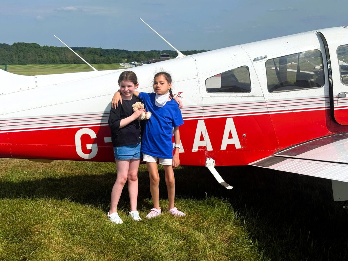 Two children stand side by side on grass with their arms around each other beside a small red and white aircraft at an aerodrome, with a runway and trees visible in the background on a sunny day.