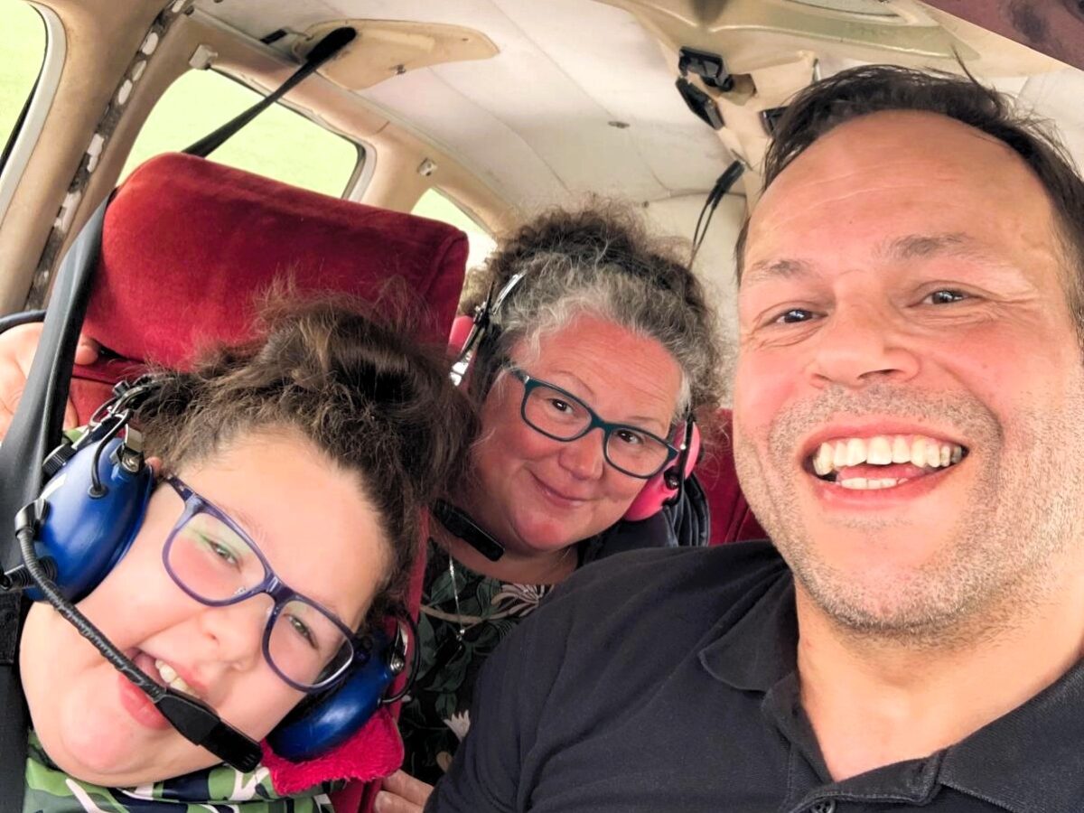 Three people sit inside a small aircraft wearing aviation headsets, posing for a photo in the cabin with red seats visible behind them.